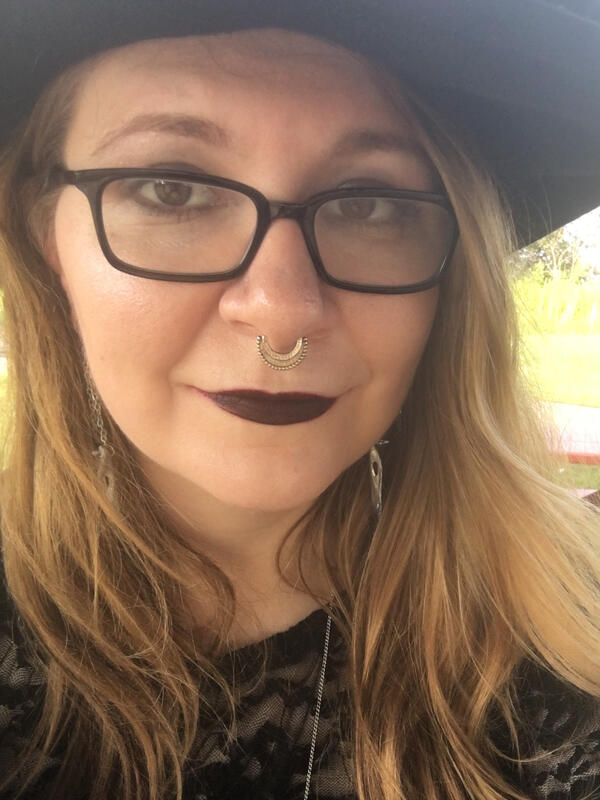 Author portrait Woman with blonde hair, dressed in black with black glasses and dark red lipstick.